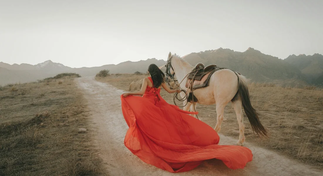Person in a flowing red dress sits on a dusty path beside a light-colored horse in a wide-open, mountainous landscape.