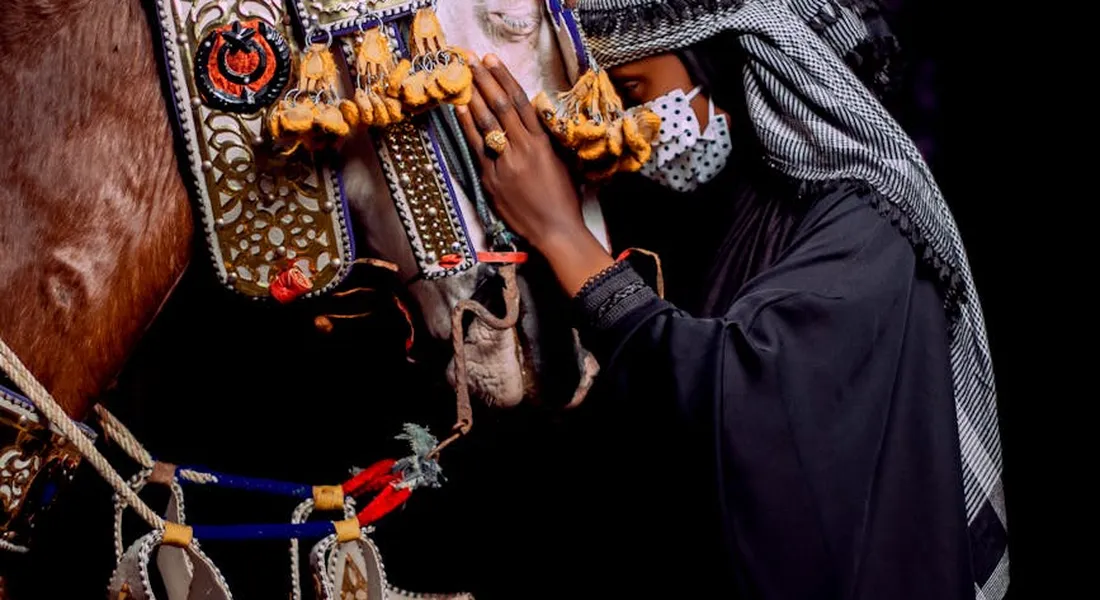 A person in dark clothing and a headscarf gently comforts a richly adorned horse, resting a hand near the horse's muzzle as it stays calm.