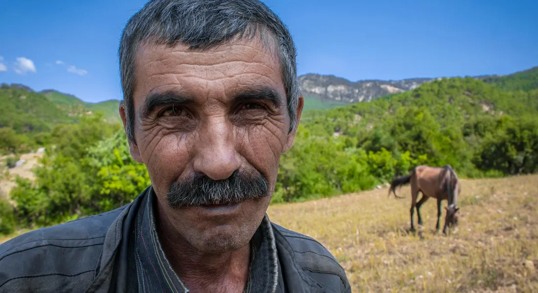 Close-up of a middle-aged man with a mustache in a sunlit pasture; a horse grazes in the background among green hills.