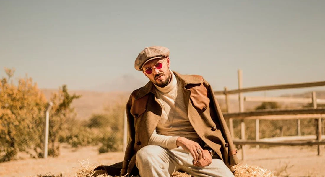 A man wearing a brown jacket, cap, and red sunglasses sits outdoors in a dry, rural landscape.