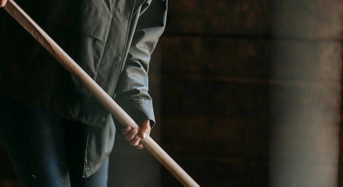 Close-up of a worker in workwear gripping a long-handled tool inside a dim stable.