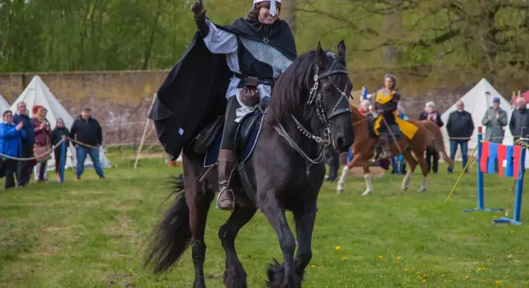 A rider wearing a cloak rides a black horse across a grassy field at an outdoor event, with tents and spectators visible in the background.