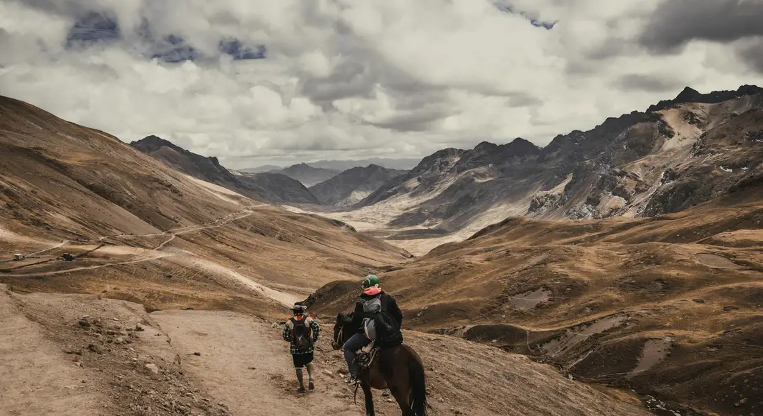 A rider on a horse along a rugged mountain trail with a hiker walking beside, set against sweeping, rocky valleys.