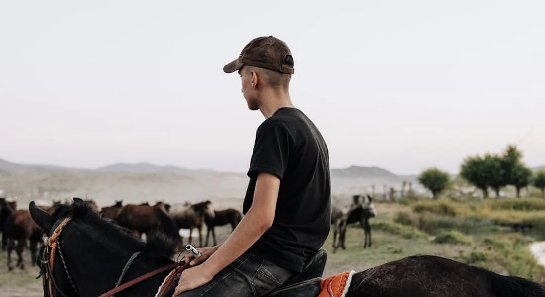 A rider on horseback wearing a black t-shirt and cap, looking toward a distant herd of horses in an open, arid landscape.