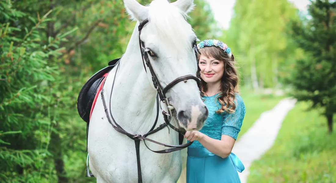 A woman wearing a blue dress and a flower crown stands beside a white horse on a grassy path, surrounded by green trees.