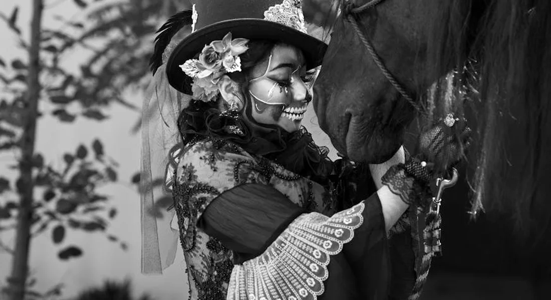 Black-and-white photo of a person wearing a decorated hat and face paint closely interacting with a dark horse, with a leafy background.