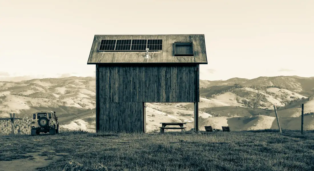 Sepia-toned photo of a small weathered wooden shelter in a grassy hillside pasture, with a bench nearby and an old vehicle on the left, mountains in the distance.