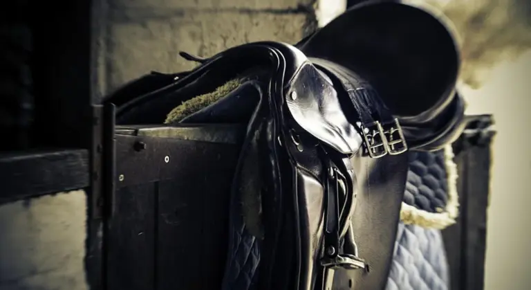 Close-up of a riding saddle and tack hanging on a rack in a stable, highlighting gear readiness