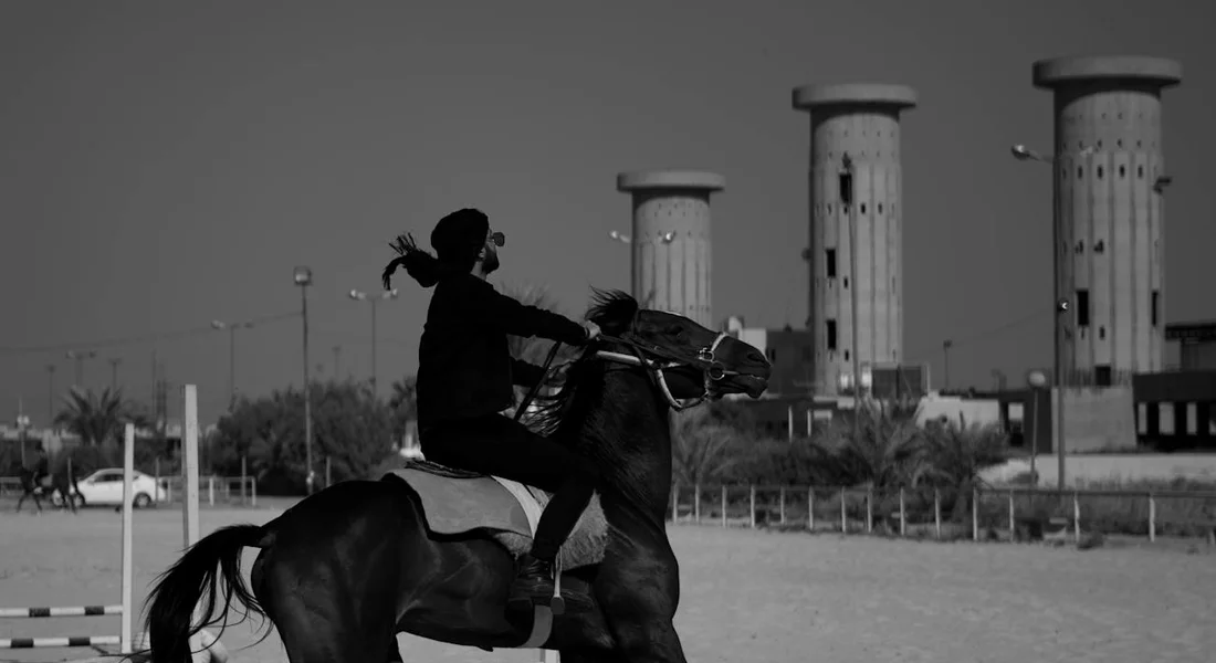 Rider on a horse mid-jump over a low obstacle in an outdoor arena; black-and-white photograph