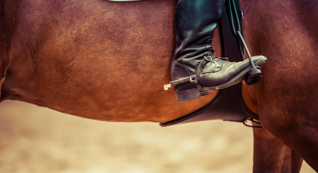 Close-up of a rider's boot in a stirrup next to a horse, with saddle and tack, on a sandy arena.