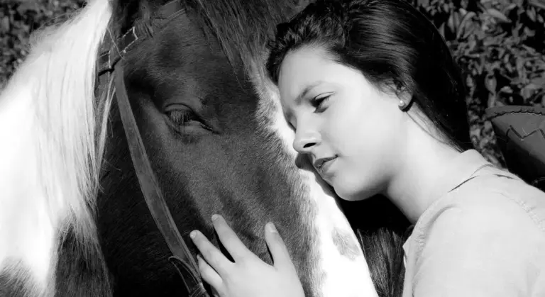 Close-up, black-and-white portrait of a person with their forehead resting gently against a horse's face, eyes closed in a tender moment.