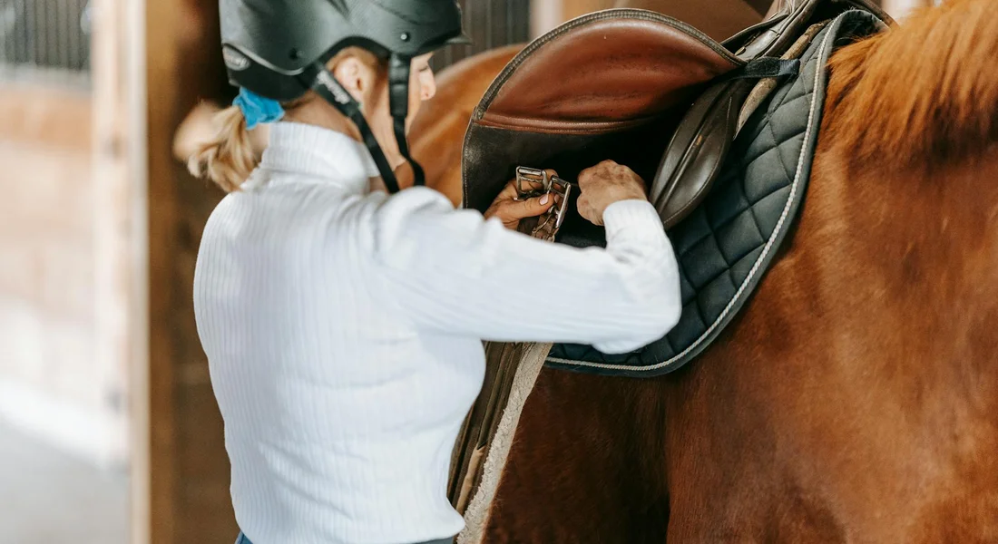 Rider wearing a helmet adjusts a saddle on a horse in a stable.