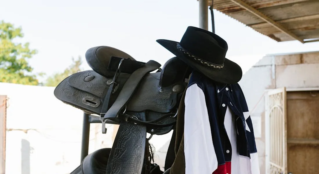 A horse saddle rests on a rack with a black cowboy hat hanging nearby, ready for use.