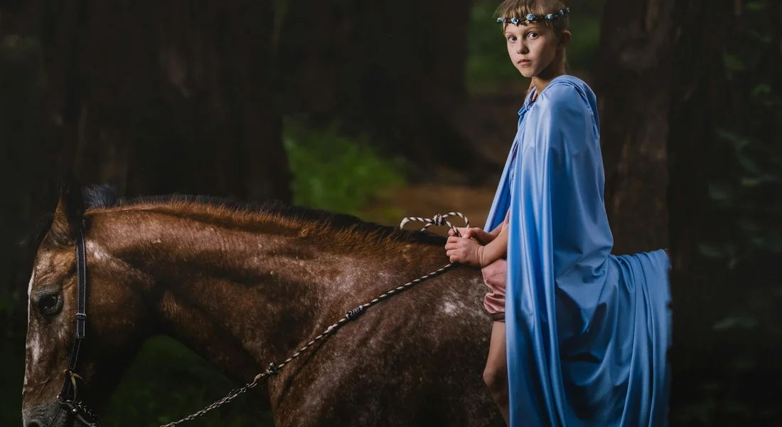 Rider on a brown horse in a forest, wearing a blue cloak and looking toward the camera.