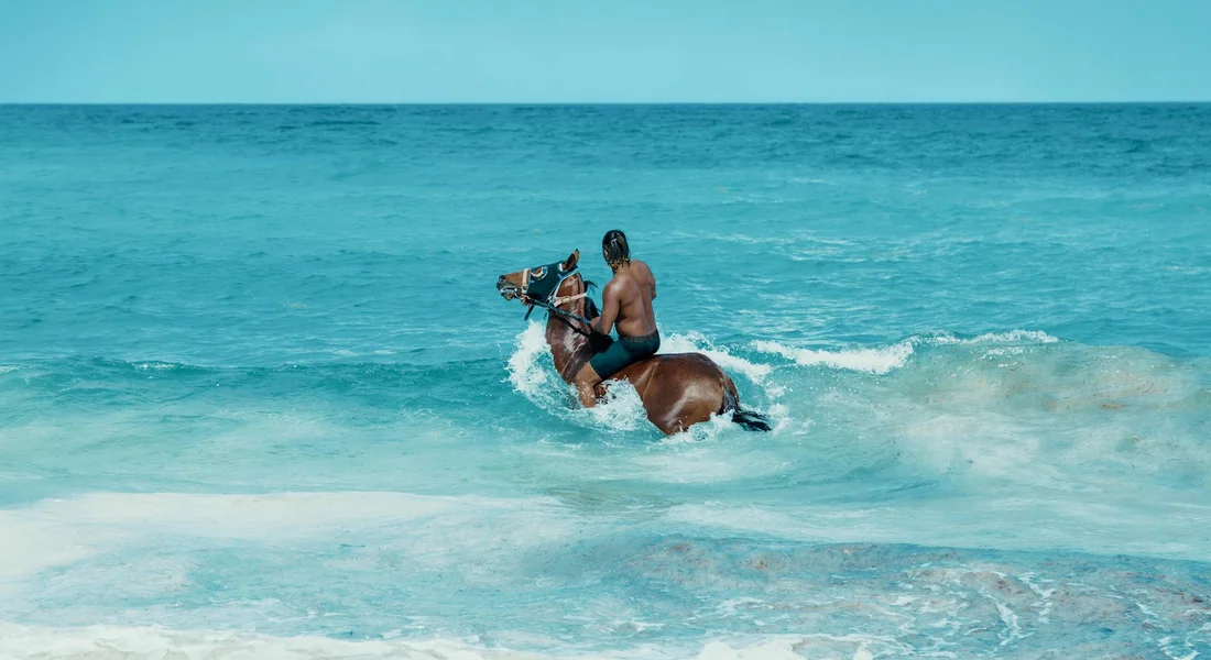 A shirtless man rides a horse through turquoise ocean waves near a sandy beach