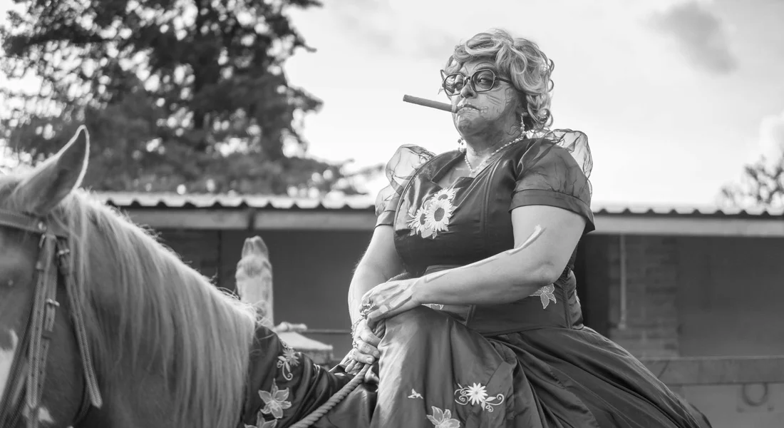 Older woman in a dress sits on a horse outdoors, smoking a pipe