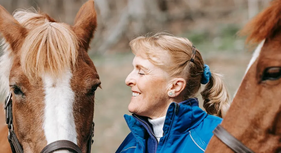 Smiling woman in a blue jacket standing between two horses, illustrating post-clean maintenance and care.