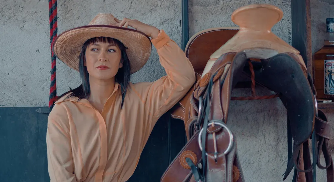 Person in a wide-brimmed hat standing beside a saddle and horse tack in a stable, ready for organized storage and inspection.