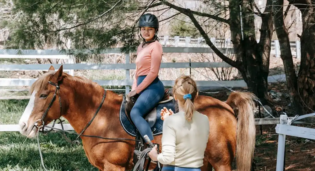 A rider in a pink top and helmet sits on a chestnut horse while a handler stands nearby, observing the horse in a shaded riding area.