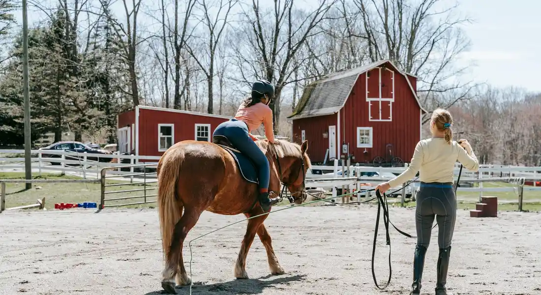 Horse and rider in a riding arena with a trainer holding the lead rope; a red barn and leafless trees in the background.