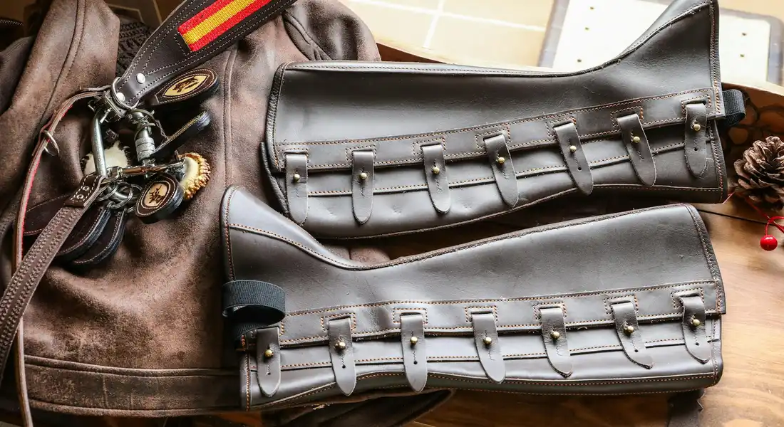 Two dark leather saddle bags with multiple buckles laid out on a table with other riding gear, illustrating tack sized for a larger horse frame.