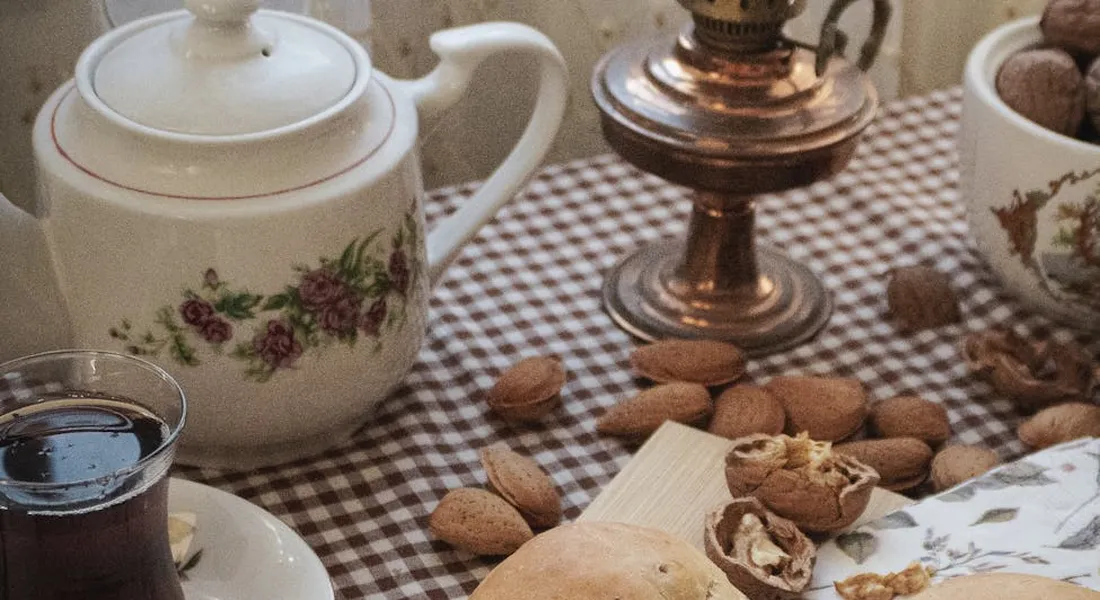 A cozy tea setting on a checkered tablecloth featuring a white porcelain teapot, a glass of dark tea, a brass lamp, and scattered almonds.