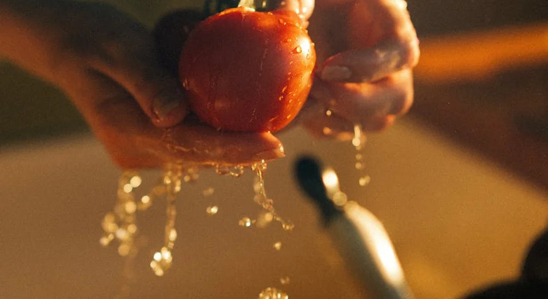 Hands rinsing a small red tomato under running water.