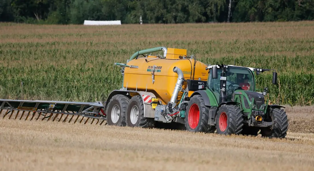 Green tractor pulling a yellow manure-spreading tanker across a harvested field
