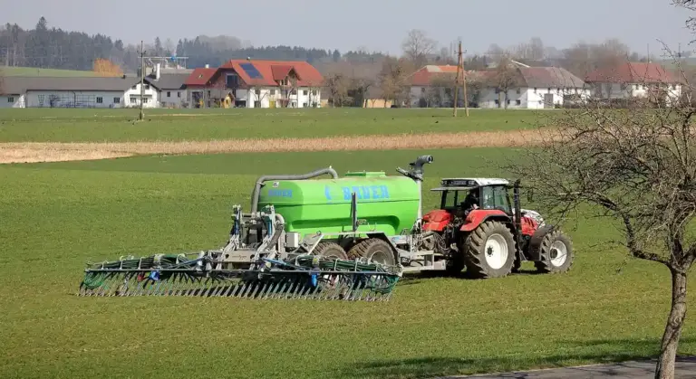 Red tractor pulling a green manure spreader across a grassy field with farm buildings in the background