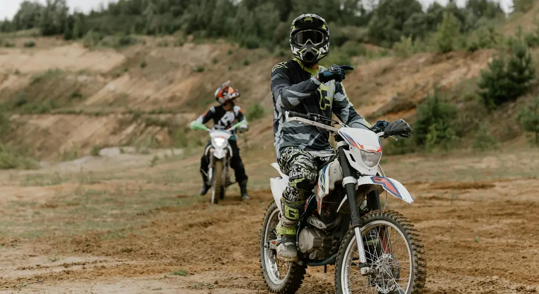 Two off-road dirt bike riders on a dry, dusty trail. The foreground rider wears protective black gear and a helmet, while a second rider follows in the background on a white bike; terrain is rugged with sparse vegetation and hills in the distance.