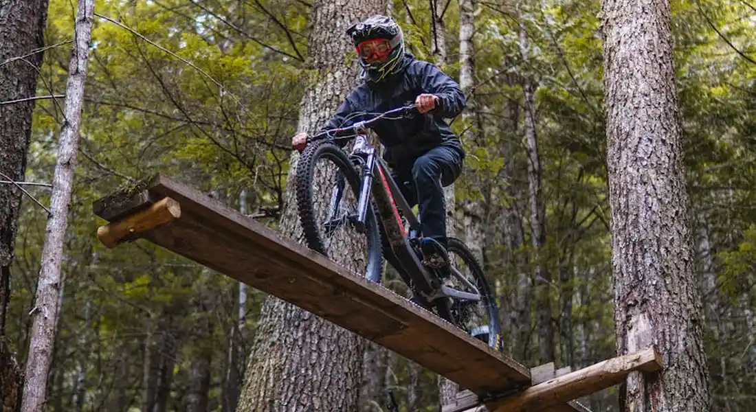 Mountain biker wearing a helmet crosses a wooden log bridge on a forest trail.
