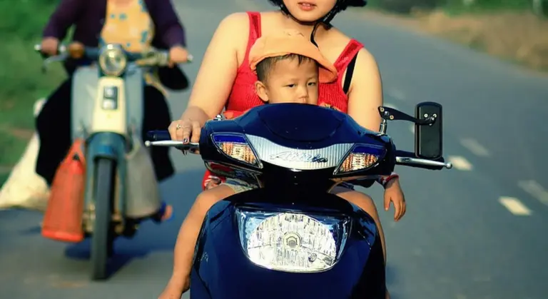 Adult rider on a blue scooter with a child passenger on the front, riding on a road with another rider on a scooter in the background