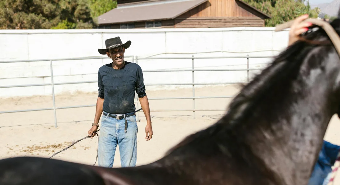 Trainer in a wide-brimmed hat and jeans stands in an outdoor arena holding a lead rope, with a dark horse's head visible in the foreground.