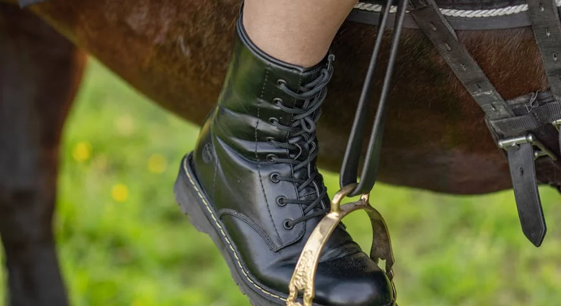 Close-up of a rider's black leather riding boot in a stirrup, with a horse's leg and saddle straps visible in the background.