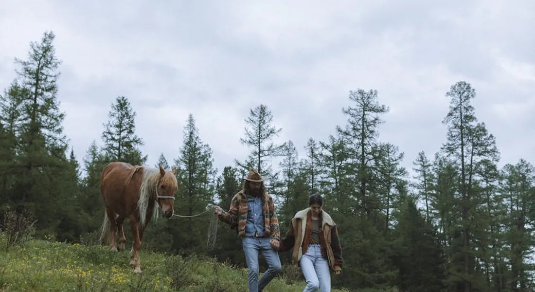 Two people walking a horse through a grassy meadow with tall trees in the background.