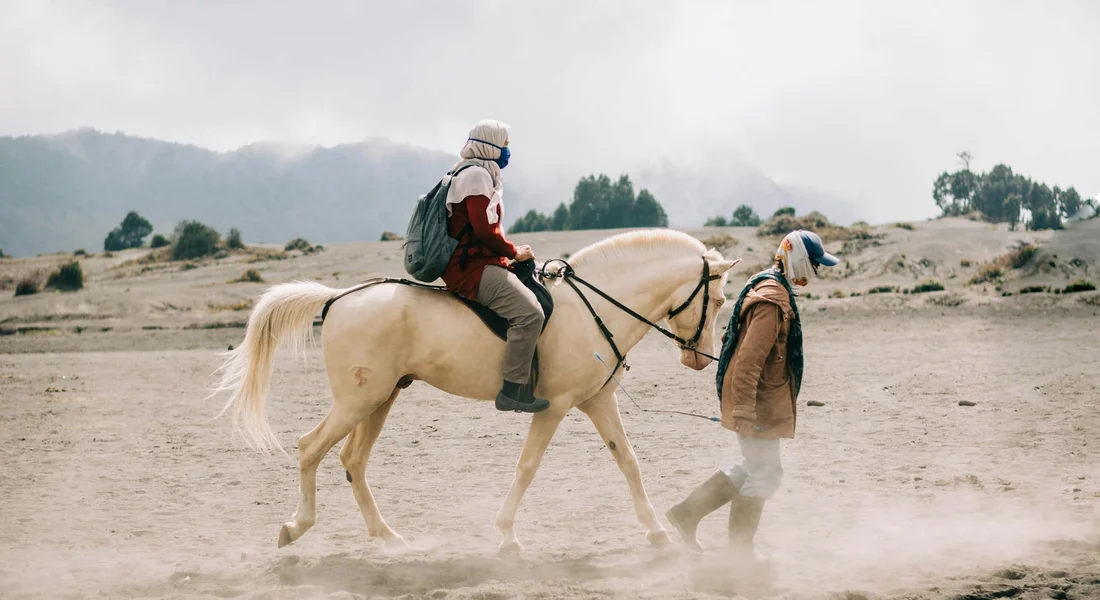 Two people in outdoor gear, one mounted on a light-colored horse and the other walking beside, in a dusty arena with distant hills.