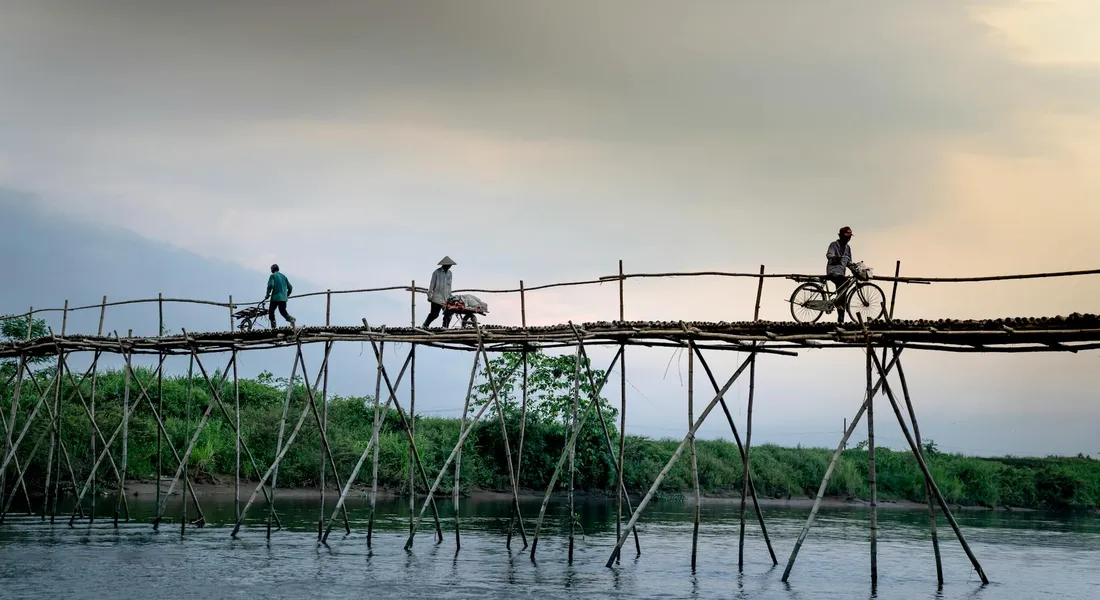 A rustic wooden bridge spans a calm river, with several people walking and a cyclist crossing toward the far end.