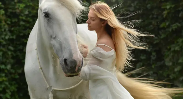 A woman in a flowing white dress stands beside a white horse in a garden.