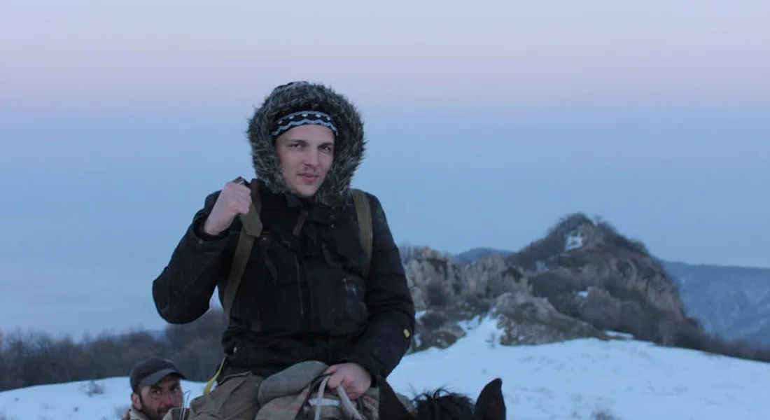 A winter rider on a horse, wearing a hooded jacket with fur trim, sits atop a snow-covered hillside with rocky outcrops in the distance.
