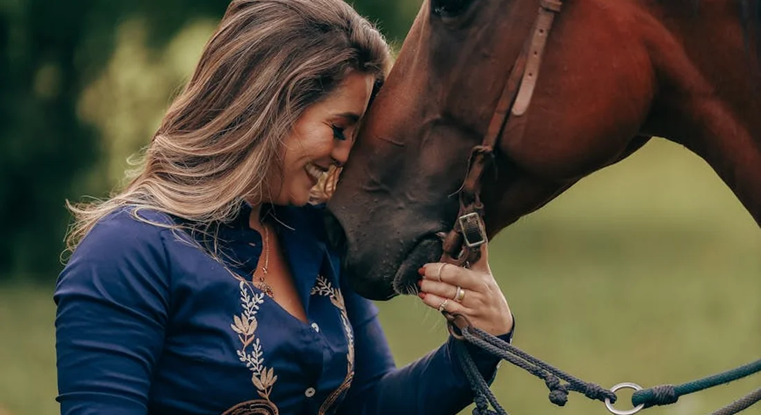 Smiling woman in a blue blouse gently touching the nose of a calm horse in a close-up.