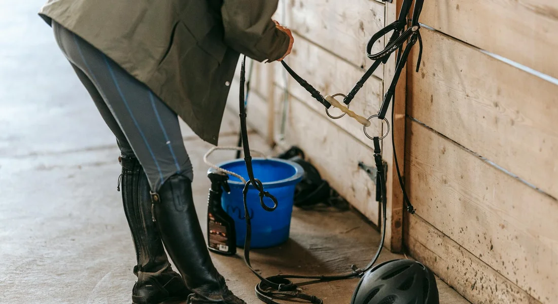 A person in riding boots and a jacket stands by a horse stall, handling tack with a blue bucket nearby and a riding helmet resting on the floor.